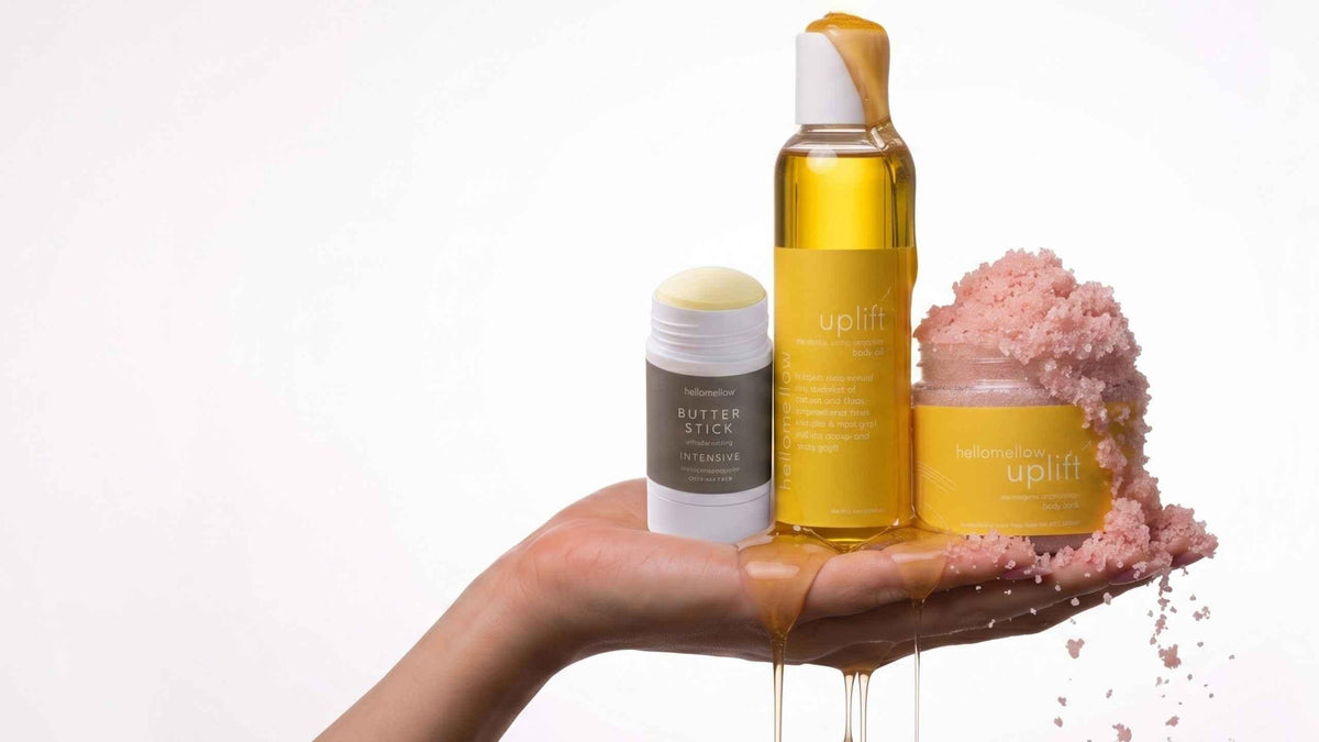 a woman's hand holding hellomellow scrub body oil and body butter stick in uplift