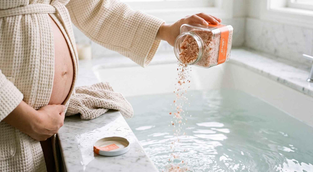 pregnant woman pouring mineral bath salts into lukewarm water beside a bright bathroom window.