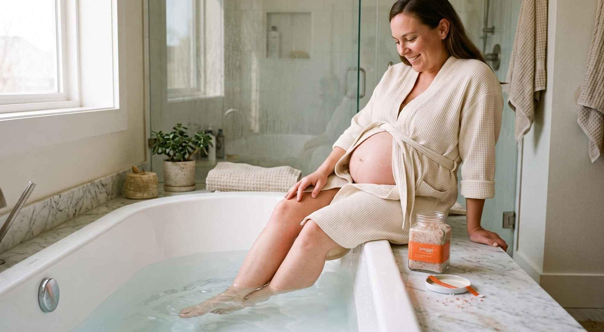 pregnant woman soaking her feet and lower legs beside mineral bath salts in a bright bathroom.