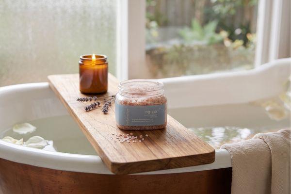 hellomellow relax bath salts sit on a wooden tray beside a warm candlelit bath.